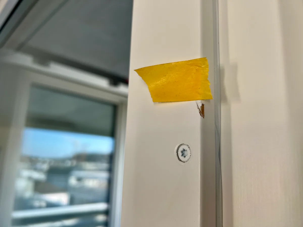 Man sieht einen gelben Klebestreifen an einem weiss bemalten Holzrahmen neben einem Fenster. Unter dem Klebestreifen ist eine kleine Delle an der Holzkante, bei der die Farbe ab ist.